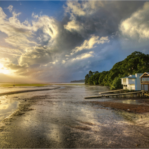 Neritic - A white and brown wooden coastal house sits beside the seashore at sunrise, glowing with warm early light as waves gently meet the shoreline.