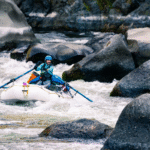 Kayaker navigating Blossom Bar rapid on the Rogue River, surrounded by rocks, representing river sports culture, photographed by Ben Kitching