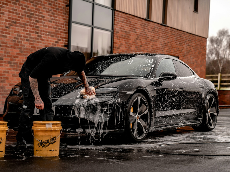 Man in black T-shirt splashing water on a black coupe while washing a car with water during daytime.
