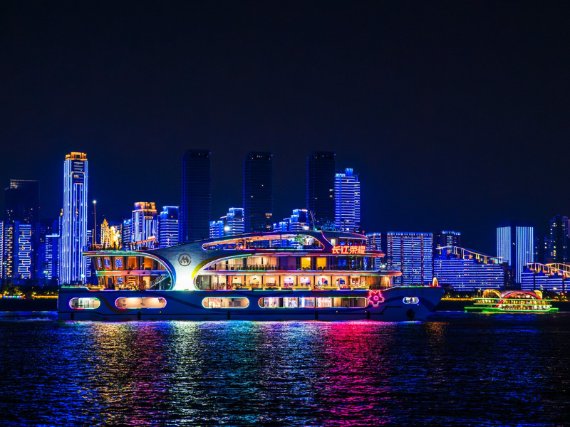 A lighted river cruise ship sailing past a glowing city skyline at night, highlighting the atmosphere of themed river cruises along urban waterways.