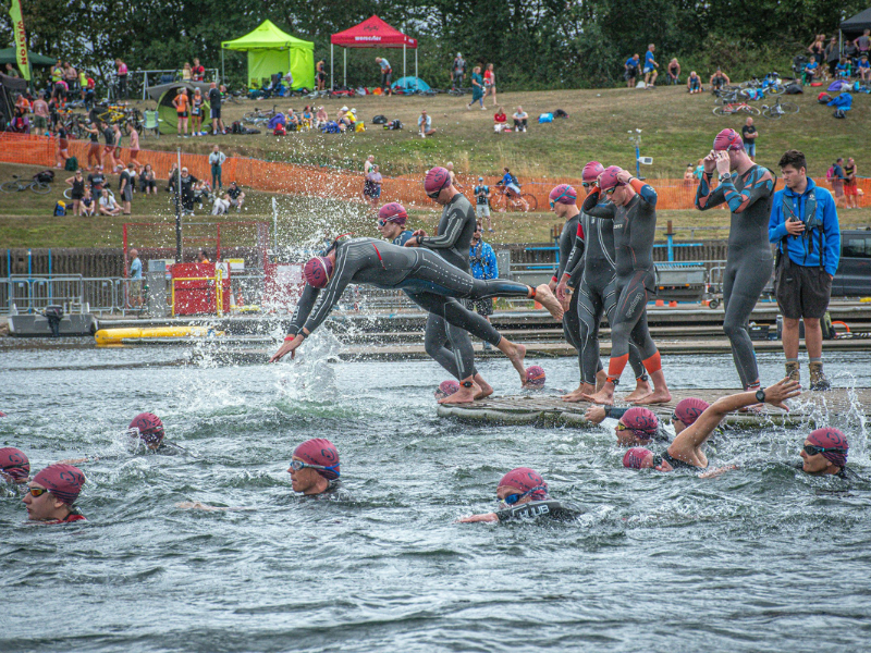 Triathletes in wetsuits begin an open water swimming race as they enter the water together at the start line
