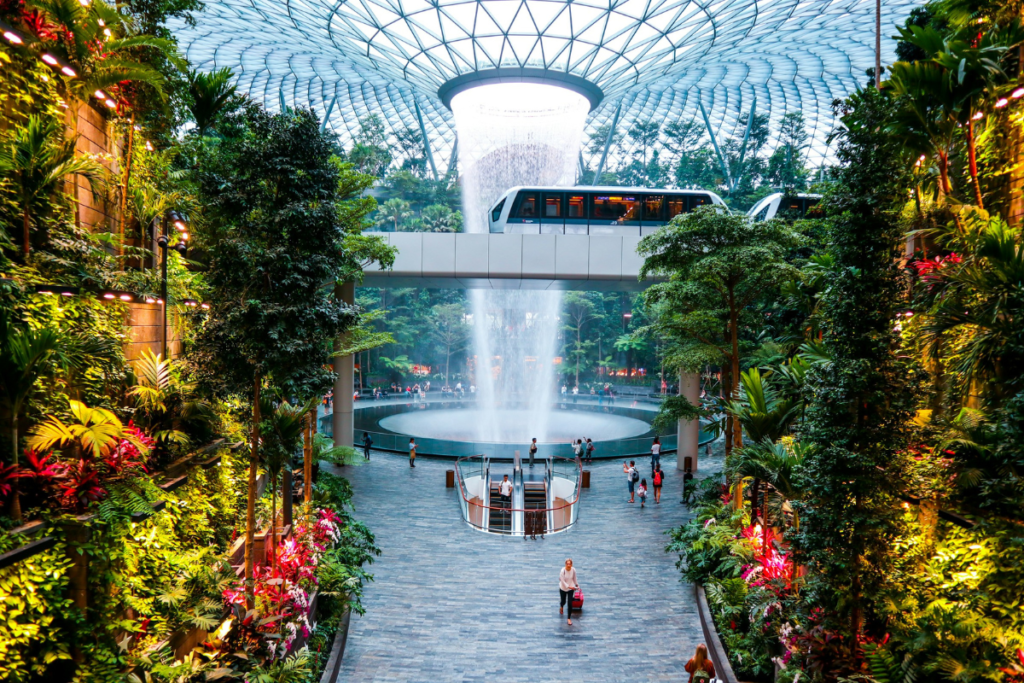 Travelers walk through the lush indoor gardens and Rain Vortex waterfall at Changi International Airport, reflecting how airports are rebranding through water and climate-focused design.