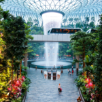 Travelers walk through the lush indoor gardens and Rain Vortex waterfall at Changi International Airport, reflecting how airports are rebranding through water and climate-focused design.