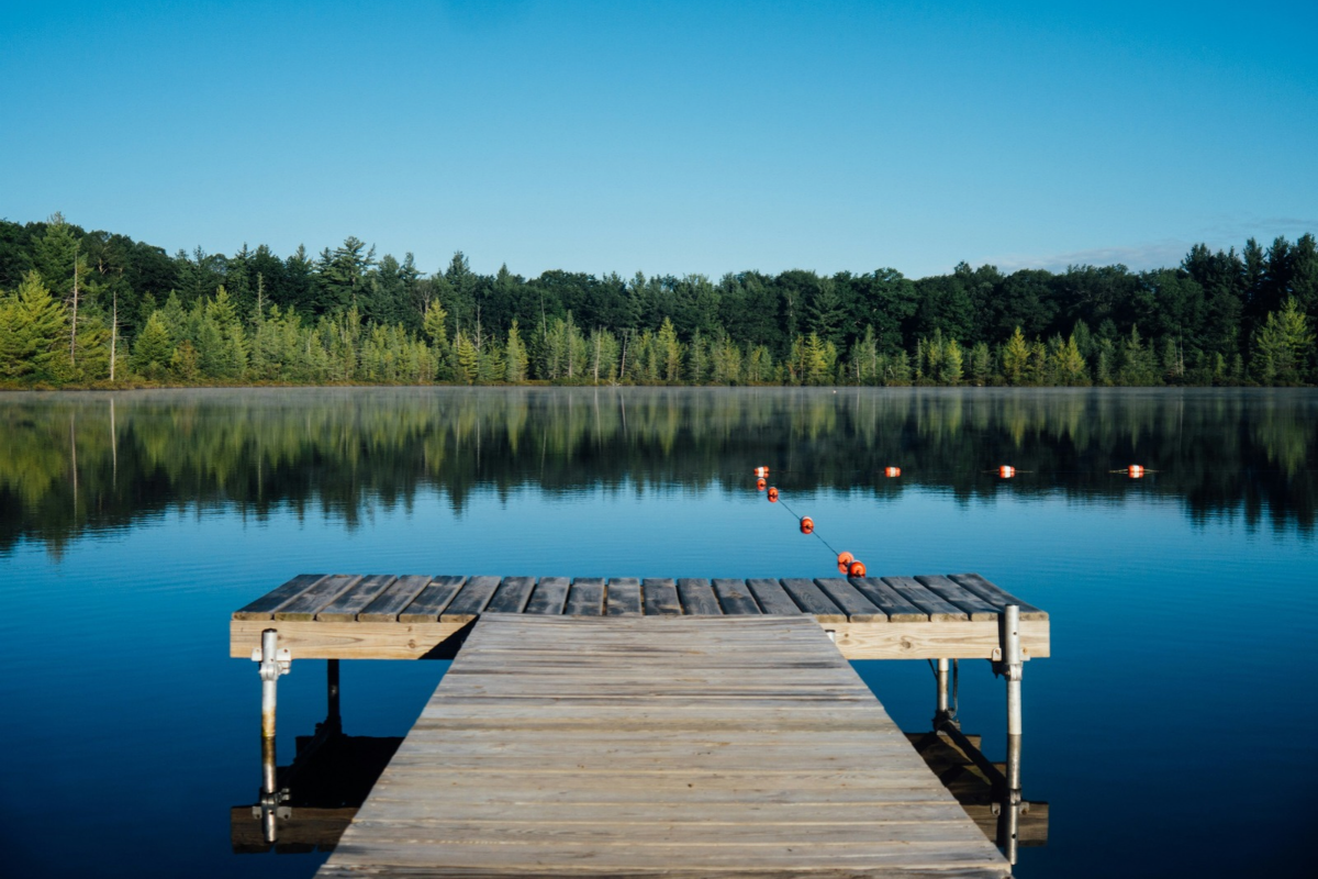 A peaceful lake with clear blue water reflecting surrounding trees, featuring a wooden dock extending towards a designated swimming area.