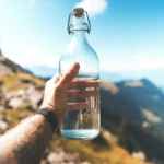 A traveller holds a glass water bottle against a mountain landscape, highlighting tourism water scarcity and the importance of freshwater conservation.
